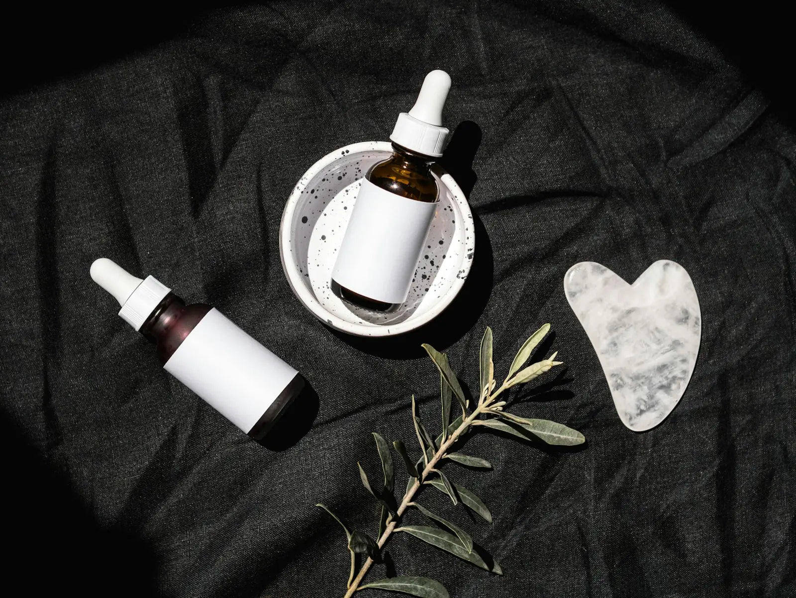 Skincare-Stillleben mit zwei Pipettenflaschen, Gua-Sha-Stein und Olivenzweig auf schwarzem Leinen